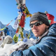 Person wearing sunglasses and winter gear sits on a snowy mountain with colorful prayer flags in the background.